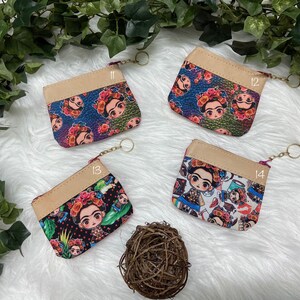 May include: Four small coin purses with zipper closures and keychains. Each purse features a different colorful Frida Kahlo design on the front, with a tan top and a gold-colored zipper. The purses are displayed on a white surface with greenery.