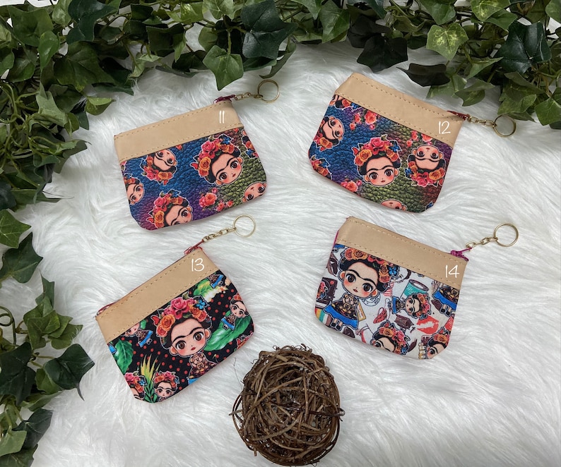 May include: Four small coin purses with zipper closures and keychains. Each purse features a different colorful Frida Kahlo design on the front, with a tan top and a gold-colored zipper. The purses are displayed on a white surface with greenery.