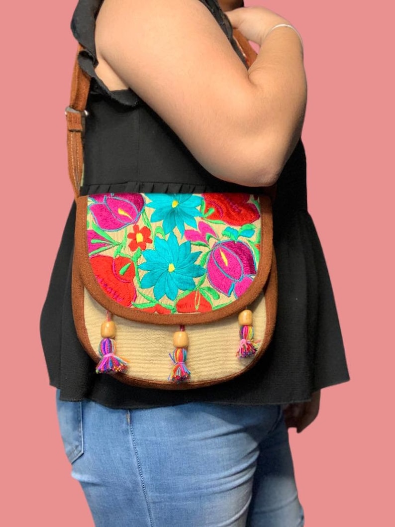May include: A brown crossbody bag with a colorful floral embroidered flap. The bag has a brown strap and wooden bead tassels.