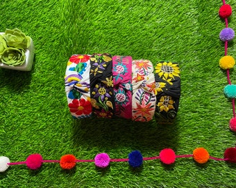 May include: Five colorful embroidered headbands with floral and geometric patterns. The headbands are made of fabric and have a knotted design. They are arranged in a stack on a green grassy surface.