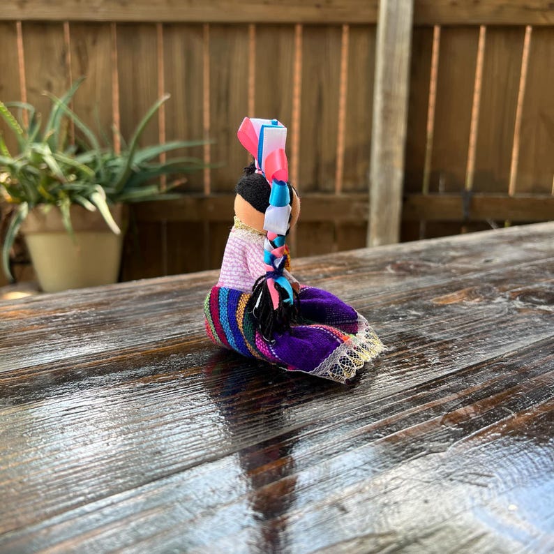 Op de afbeelding: Een kleine handgemaakte pop met een kleurrijke rok en een paars bovenstuk. De pop heeft zwart haar met een blauw en roze lint dat erin is gebonden.