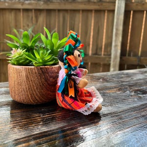Op de afbeelding: Een kleurrijke handgemaakte pop met een rode, oranje en groene rok, een roze blouse en een groen, oranje en blauw lint hoofdband. De pop zit op een houten oppervlak.