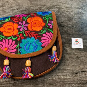 May include: A brown canvas crossbody bag with colorful floral embroidery and brown tassels. The bag has a brown suede trim and wooden beads.