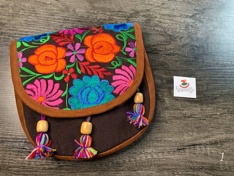 May include: A brown canvas crossbody bag with colorful floral embroidery and brown tassels. The bag has a brown suede trim and wooden beads.