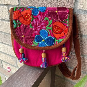 May include: A pink fabric crossbody bag with a brown suede flap. The flap is embroidered with colorful flowers in shades of red, orange, blue, green, and purple. The bag has wooden beads and colorful tassels.