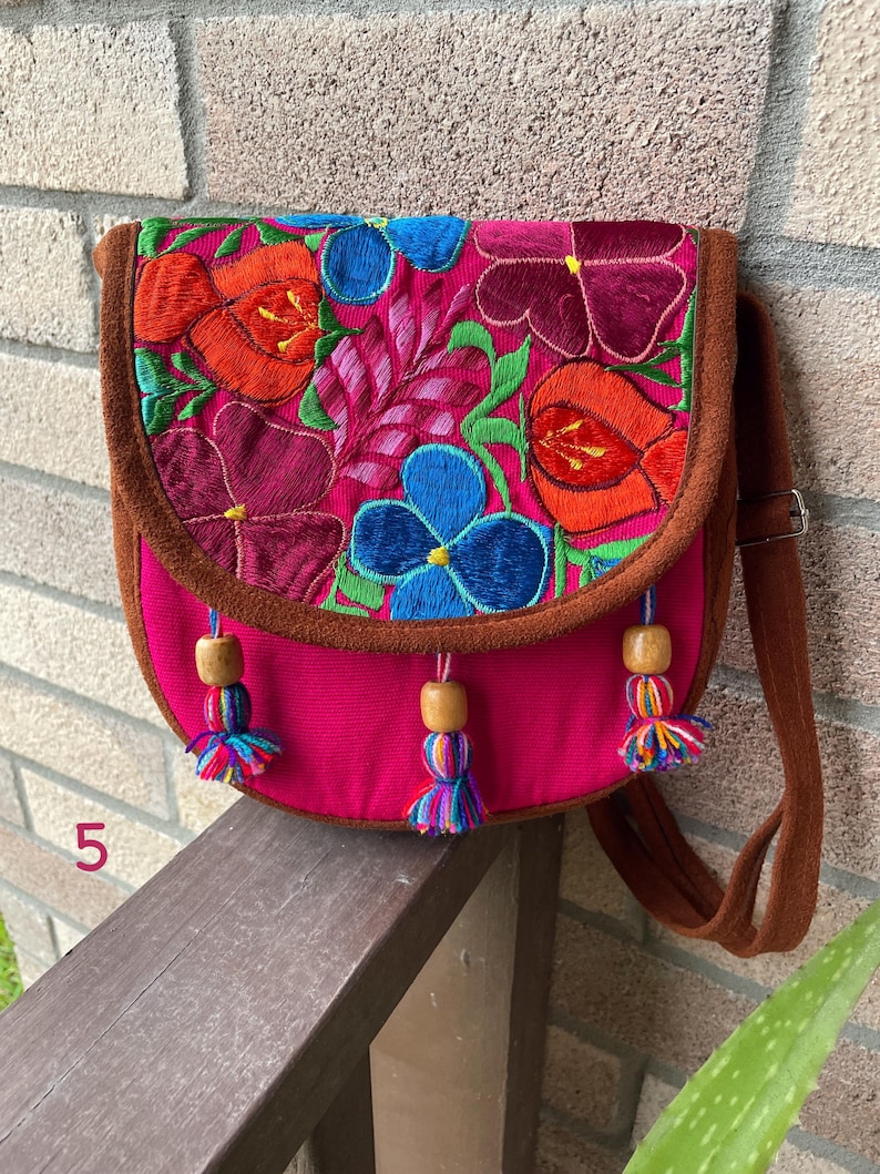 May include: A pink fabric crossbody bag with a brown suede flap. The flap is embroidered with colorful flowers in shades of red, orange, blue, green, and purple. The bag has wooden beads and colorful tassels.