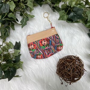 May include: A small, rectangular coin purse with a beige top and a colorful patterned bottom featuring a portrait of Frida Kahlo. The purse has a gold-colored keychain and the number "17" printed on it. The background is white with green foliage.