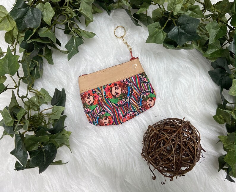 May include: A small, rectangular coin purse with a beige top and a colorful patterned bottom featuring a portrait of Frida Kahlo. The purse has a gold-colored keychain and the number "17" printed on it. The background is white with green foliage.