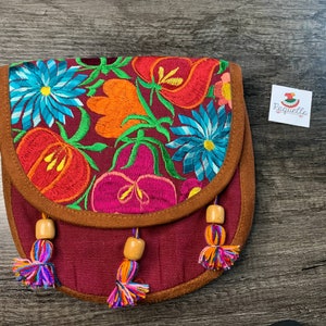 May include: A small, burgundy fabric purse with a brown suede flap and colorful floral embroidery. The purse has wooden beads and tassels.