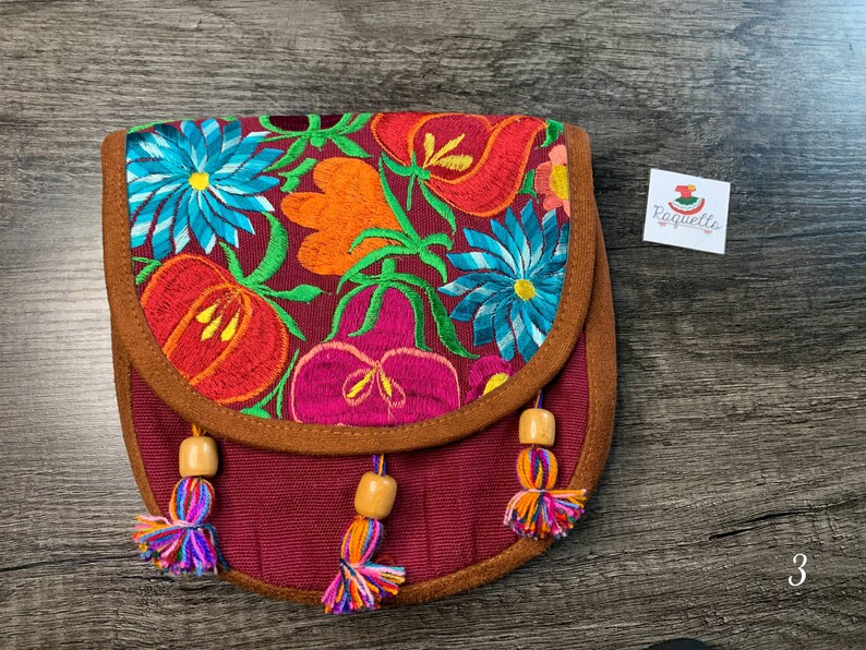 May include: A small, burgundy fabric purse with a brown suede flap and colorful floral embroidery. The purse has wooden beads and tassels.