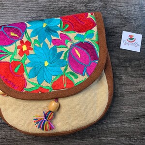 May include: A small beige fabric purse with a brown trim and a floral embroidery design in shades of pink, red, blue, and green. The purse has a wooden bead tassel.