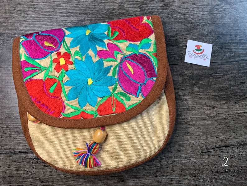 May include: A small beige fabric purse with a brown trim and a floral embroidery design in shades of pink, red, blue, and green. The purse has a wooden bead tassel.