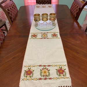 May include: A white table runner with embroidered floral designs in shades of red, yellow, and green. The runner has a fringe border and is displayed on a wooden table with three candle holders.