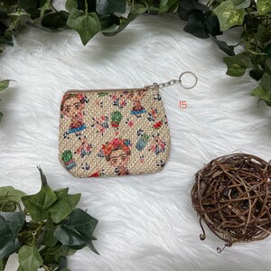 May include: Small beige coin purse with a zipper closure and a key ring. The purse is decorated with a repeating pattern of a woman's portrait, flowers, and cacti. The background is a soft, white, textured surface.
