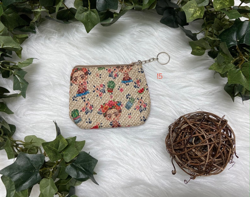 May include: Small beige coin purse with a zipper closure and a key ring. The purse is decorated with a repeating pattern of a woman's portrait, flowers, and cacti. The background is a soft, white, textured surface.