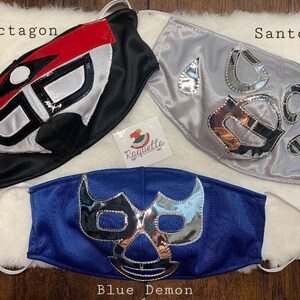 K&ouml;nnte beinhalten: Drei Wrestling-Masken, eine schwarze mit roten und wei&szlig;en Akzenten, eine silberne mit silbernen Akzenten und eine blaue mit silbernen Akzenten. Die Masken sind mit "Octagon", "Santo" und "Blue Demon" beschriftet.