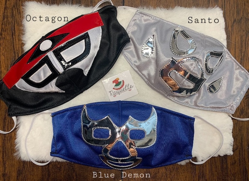 K&ouml;nnte beinhalten: Drei Wrestling-Masken, eine schwarze mit roten und wei&szlig;en Akzenten, eine silberne mit silbernen Akzenten und eine blaue mit silbernen Akzenten. Die Masken sind mit "Octagon", "Santo" und "Blue Demon" beschriftet.