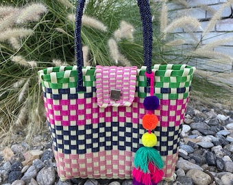 May include: A colorful woven tote bag with a rectangular shape. The bag features a woven pattern in pink, green, navy, and light pink. It has navy blue handles and a matching clasp. A tassel with multiple colored pom-poms hangs from the side.
