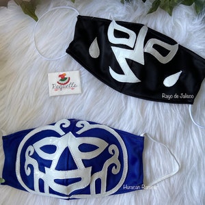 K&ouml;nnte beinhalten: Zwei Luchador-Wrestling-Masken, eine schwarze mit wei&szlig;en Akzenten und dem Text "Rayo de Jalisco" und eine blaue mit wei&szlig;en Akzenten und dem Text "Huracan Ramirez".
