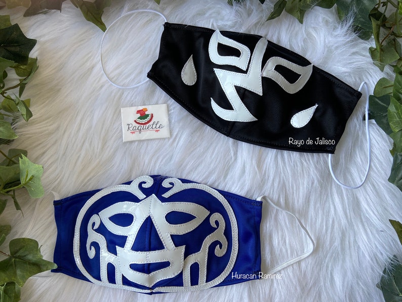 K&ouml;nnte beinhalten: Zwei Luchador-Wrestling-Masken, eine schwarze mit wei&szlig;en Akzenten und dem Text "Rayo de Jalisco" und eine blaue mit wei&szlig;en Akzenten und dem Text "Huracan Ramirez".