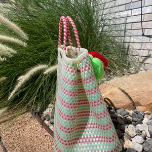 May include: A green and pink striped tote bag with two handles and a green and red tassel. The bag is made of woven plastic.