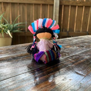 Op de afbeelding: Een kleine handgemaakte pop met zwart haar, een roze-paarse jurk en een kleurrijk lint hoofdband.