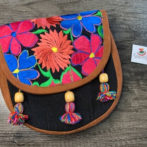 May include: A black fabric purse with a brown suede trim and colorful floral embroidery. The purse has three wooden bead tassels with colorful yarn.