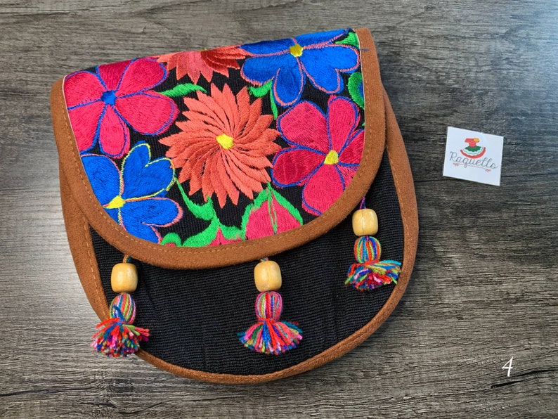 May include: A black fabric purse with a brown suede trim and colorful floral embroidery. The purse has three wooden bead tassels with colorful yarn.