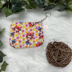 May include: A small, rectangular coin purse with a zipper closure and a silver keychain. The purse features a repeating pattern of a cartoon Frida Kahlo figure and colorful flowers on a white background. The number "16" is printed on the background.