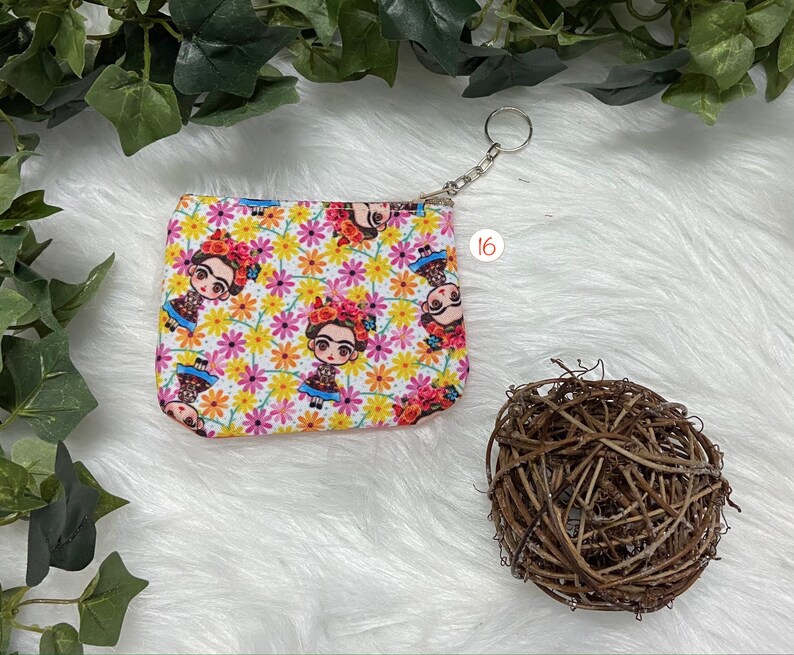 May include: A small, rectangular coin purse with a zipper closure and a silver keychain. The purse features a repeating pattern of a cartoon Frida Kahlo figure and colorful flowers on a white background. The number "16" is printed on the background.