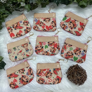 May include: Eight small, square coin purses with a Frida Kahlo design. The purses have a beige top and a zipper closure. The main body of the purses features a burlap-like fabric with a colorful Frida Kahlo print, including watermelons and flowers.