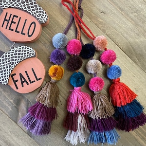 May include: A set of seven colorful pom-pom tassels with wooden acorn ornaments that say "Hello" and "Fall".