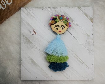 May include: A colorful embroidered felt keychain with a blue, green, and teal tassel. The keychain features a smiling face with a unibrow and a floral crown.