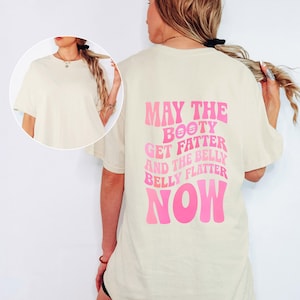 Puede incluir: Camiseta color crema con el texto rosa "MAY THE BOOTY GET FATTER AND THE BELLY BELLY FLATTER NOW". La camiseta tiene mangas cortas y un corte holgado.