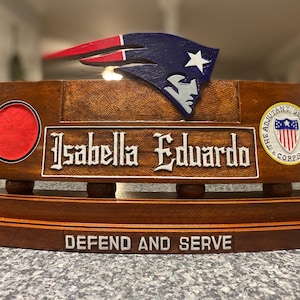 Pode incluir: Uma placa de mesa de madeira com o nome "Isabella Eduardo" em letras ornamentadas. A placa apresenta um círculo vermelho, um emblema militar e um logotipo dos Patriots. A frase "DEFEND AND SERVE" está na base.
