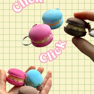 May include: A collection of colorful macaron keychains in pink, blue, and brown, each with a cream-colored filling. The keychains are displayed against a light green grid background with the word "Click" in pink.