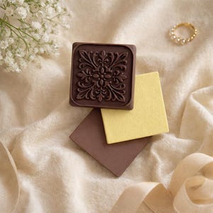 May include: A dark brown, square-shaped decorative element with an ornate floral design, resting on a light beige surface. A yellow and brown square are beneath it. A gold ring with pearls is in the background.