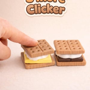May include: Two miniature s'more clickers, one with yellow filling and the other with brown filling, sit side-by-side. The top graham cracker of the s'more on the left is being lifted by a finger. The text "S'more Clicker" is above the s'mores.