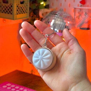 May include: A round, white dumpling keychain with a silver keyring. The dumpling has a detailed, textured design and is set in a light brown frame. The keychain is held in a hand against an orange background.