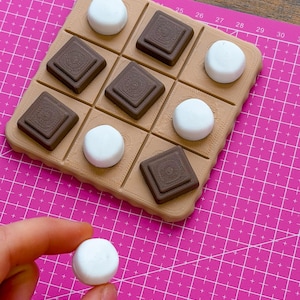 May include: A handmade tic-tac-toe game with a tan board and chocolate and marshmallow-shaped game pieces. The game pieces are brown and white. The board is on a pink cutting mat.