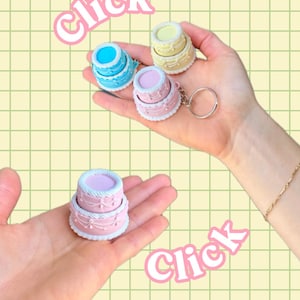 May include: Miniature cake keyrings in blue, pink, and yellow with white frosting details. The keyrings are held in a person's hands against a light green grid background. The word "Click" is written in pink.