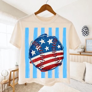 Peut inclure: T-shirt crème avec un motif de baseball. Le graphisme présente un drapeau américain vieilli sur la balle, avec des rayures rouges et blanches, des étoiles bleues et des coutures rouges. Des rayures verticales bleu clair sont derrière la balle.