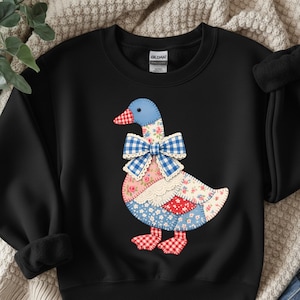 Peut inclure: Sweat-shirt noir avec un motif d'oie en patchwork. L'oie a une tête bleue, un bec à carreaux rouges et blancs et un nœud papillon en vichy bleu. Le corps est fait de tissu floral et à motifs.