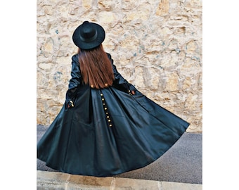 May include: A black leather coat with a long, flowing skirt and gold buttons. The coat has a wide collar and long sleeves. The model is wearing a black hat.