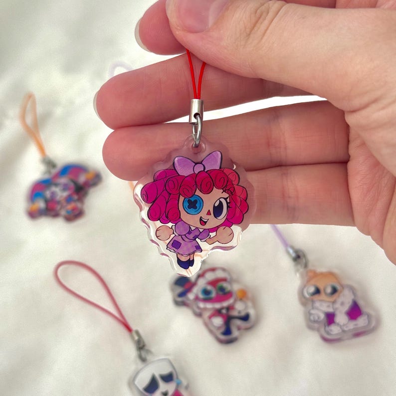May include: A clear acrylic charm featuring a cartoon girl with pink curly hair, a purple dress, and a blue eye. The charm is held by a red string. Other charms with cartoon characters are in the background.