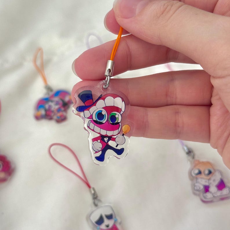May include: A clear acrylic charm featuring a cartoon character with a top hat, bow tie, and lollipop. The charm is attached to an orange lanyard. Other charms with different designs are visible in the background.