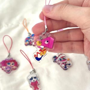 May include: A collection of colorful acrylic charms with cartoon character designs. One charm is held by a hand, featuring a pink robot-like figure. Other charms include various characters with pink, blue, and red accents, each with a matching lanyard.