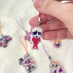 May include: Clear acrylic charm featuring a cartoon character with a white head, blue tears, and a pink outfit. The charm is attached to a red cord. Other charms with different cartoon characters are visible.