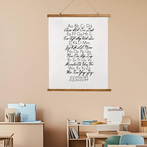 May include: A white canvas print with a cursive alphabet and numbers, displayed on a wooden frame. The print is suspended by a natural twine cord. The background is a classroom setting with desks, bookshelves, and a neutral wall.