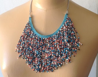 May include: A beaded necklace with a turquoise blue collar and a fringe of colorful beads in red, white, black, and blue. The necklace is displayed on a mannequin.